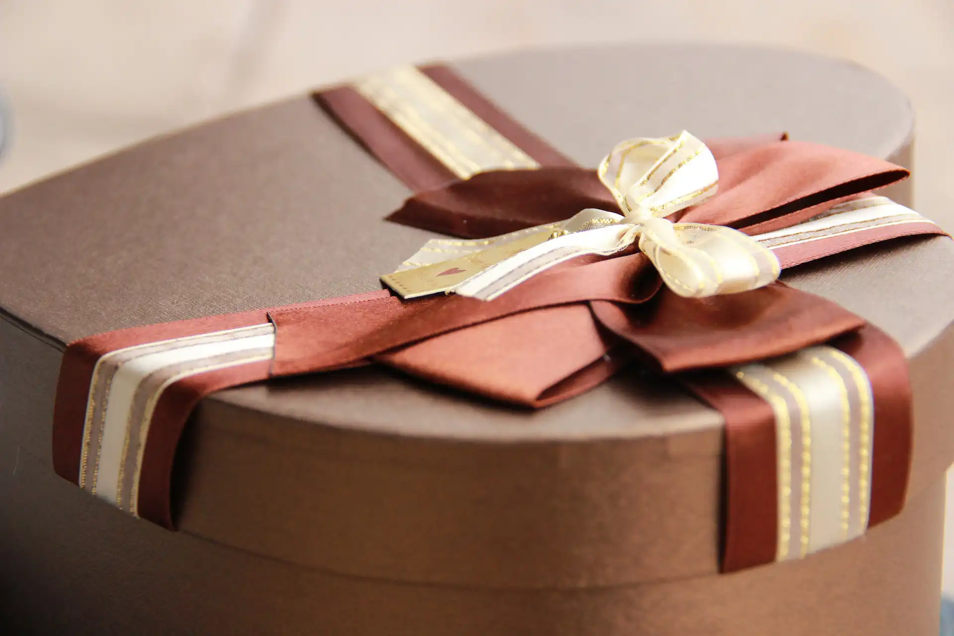 Nell'immagine c'è un pacco regalo a forma di cuore color marrone. Sopra il coperchio c'è un nastro color marrone, beige e oro che forma un grande fiocco al centro