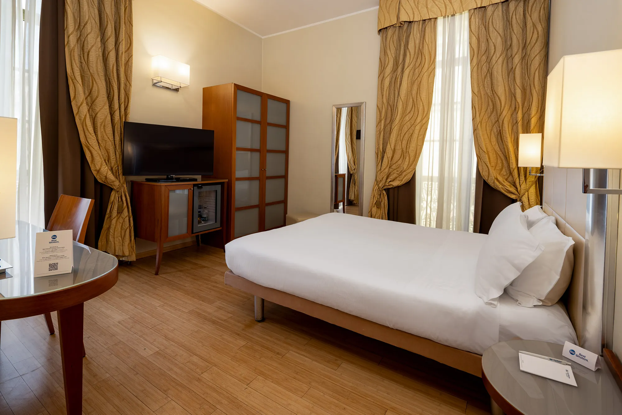 In the picture the standard double room of the hotel with the double bed on the right. Next to it is a window covered with curtains and a full-length mirror. On the left is a desk with a chair. In front of the bed is a window with curtains, a cabinet with a minibar and above it the TV. Next to it is a wardrobe. The floor is parquet..