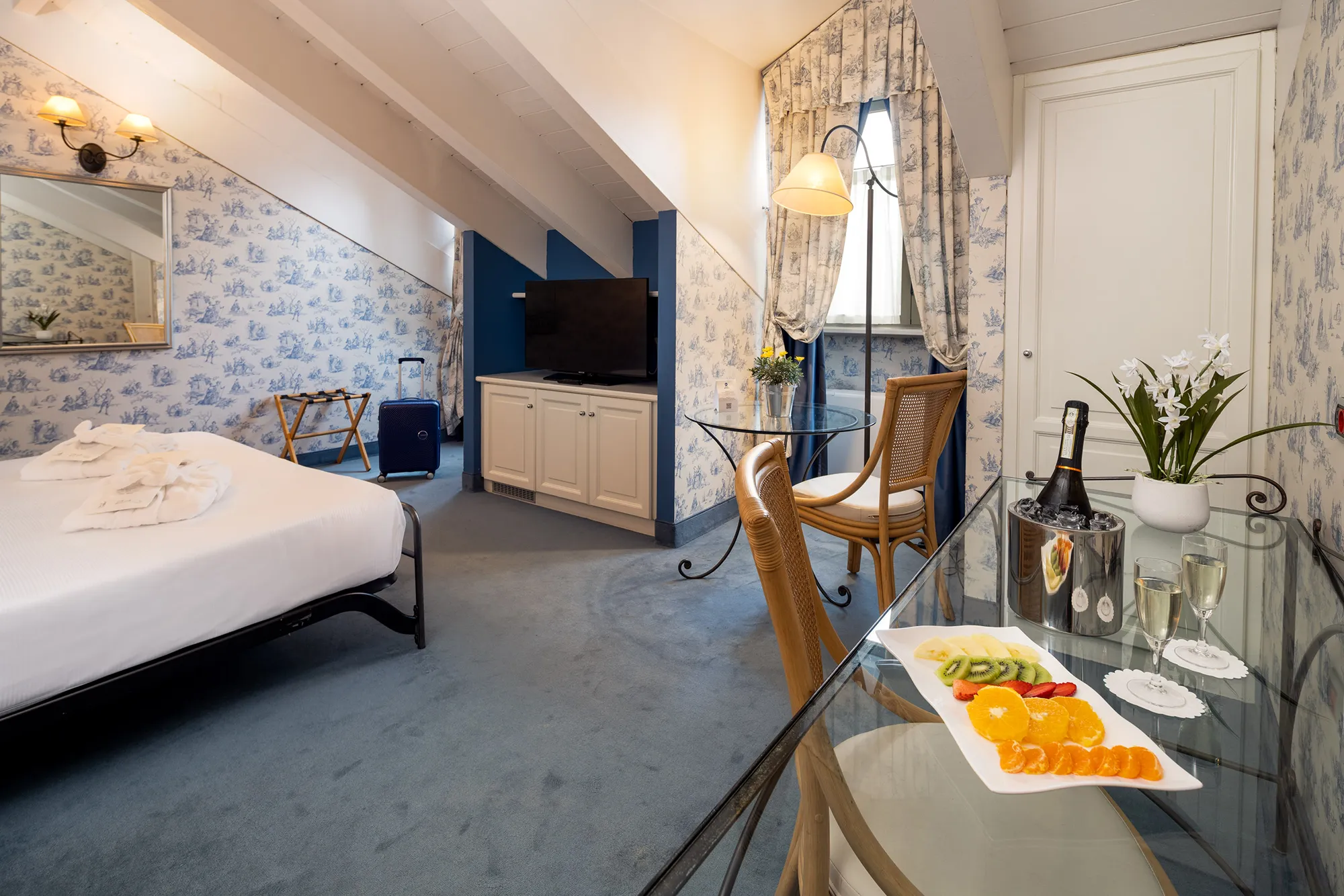 In the picture the deluxe room of the hotel. In the foreground there is a desk with a glass top and wrought iron legs and a wooden chair. On it there is a tray with some fruit in pieces, two glasses of white wine, an ice bucket with a bottle and a small plant inside. Behind it there is a closed door. On the left there is a small round table made like the desk and chair. Behind it there is a floor lamp and a window covered by curtains. To the left of the table there is a white cabinet with a TV on it. and in front of it the double bed with two bathrobes folded on top. The walls are covered with wallpaper with blue patterns and the ceiling is white attic.