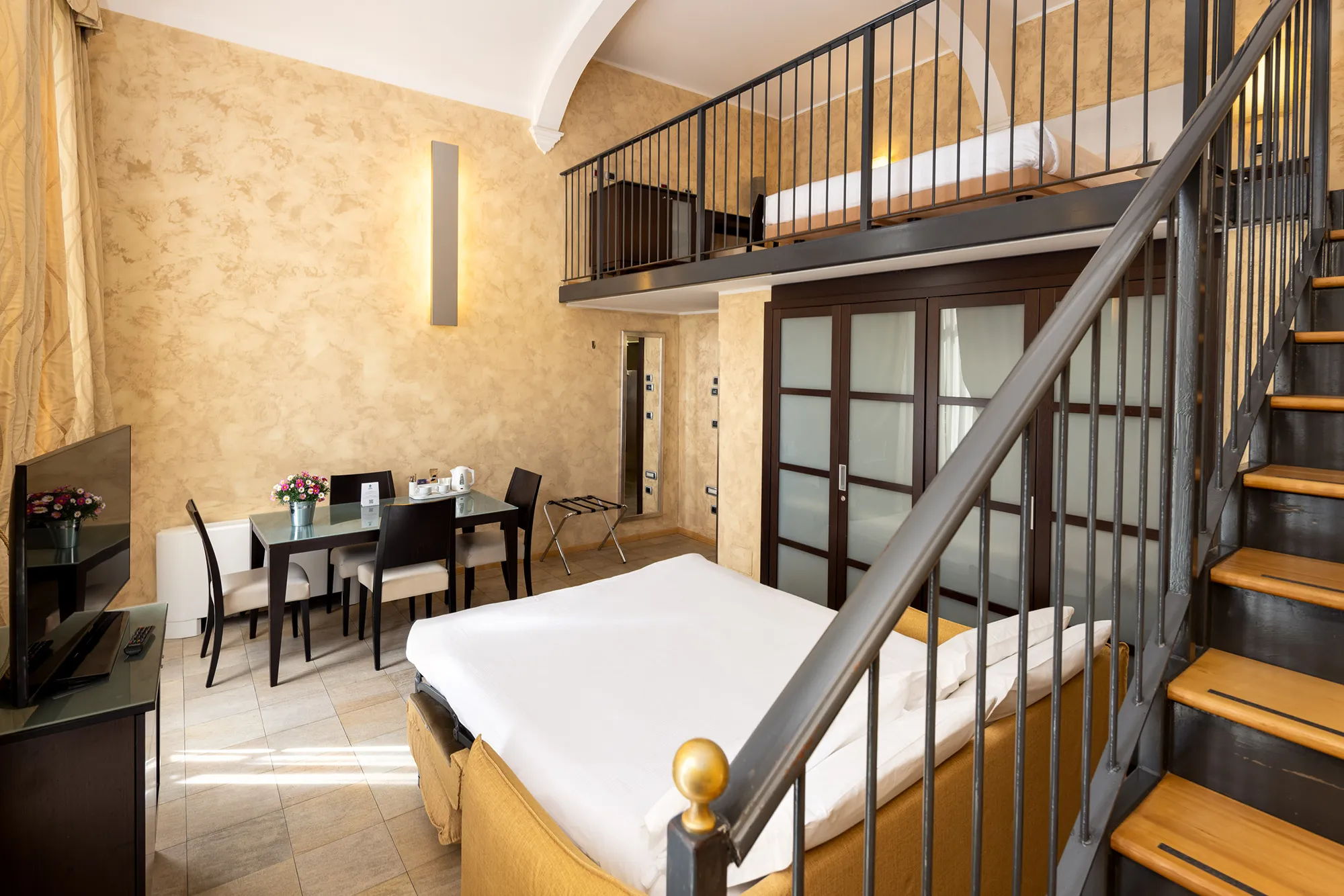 The image shows the hotel's family suite with the stairs to the upper mezzanine level in the foreground on the right, where there is a bed, a desk and a black fence. Downstairs, next to the stairs, there is an open sofa bed and on the left a cabinet with a TV on it. On the left are sliding glass and wood doors. In the center of the room is a rectangular table with chairs.