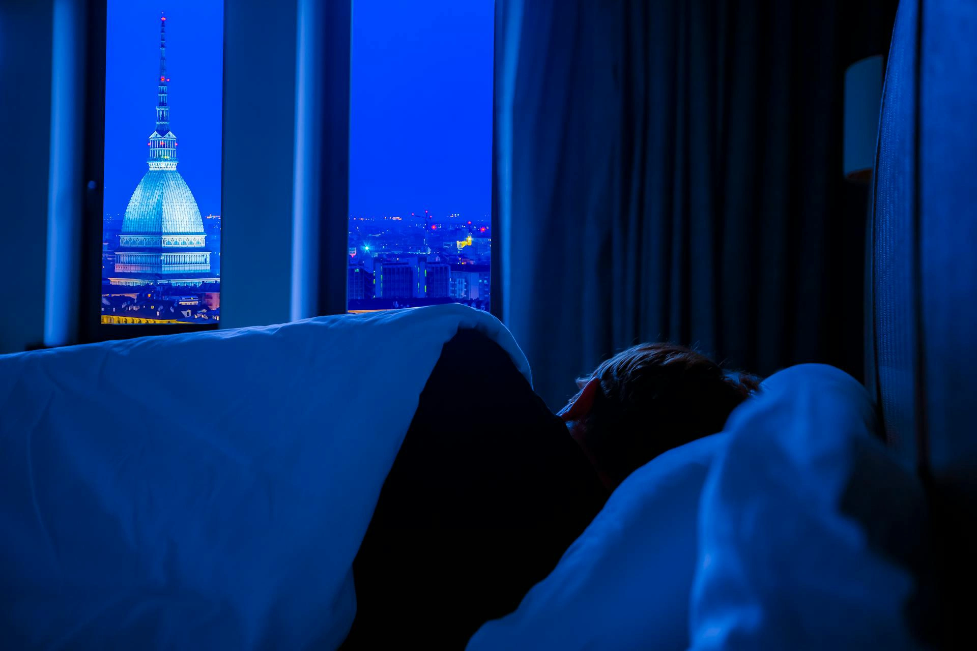 Nell'immagine si vede un uomo sdraiato a letto che dorme con il viso verso la finestra. Oltre la finestra si intravede Torino di notte e la mole antonelliana illuminata.